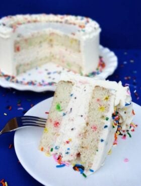 A slice of Funfetti Ice Cream Cake on white plate with the rest of the cake behind it