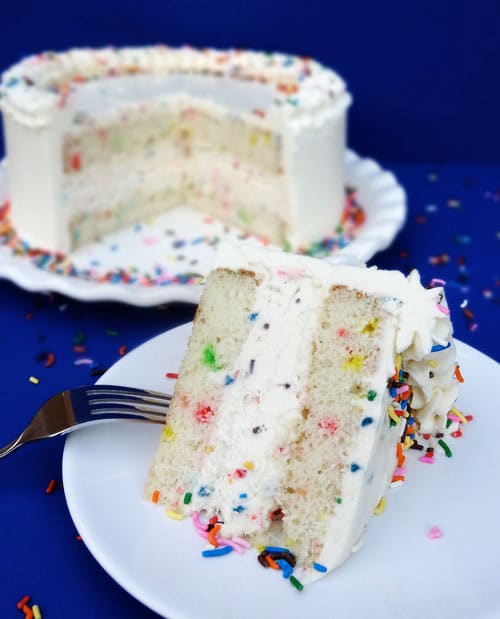 A slice of Funfetti Ice Cream Cake on white plate with the rest of the cake behind it