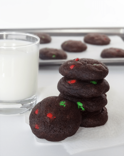 Chocolate Peanut Butter M&M Cookies are stacked next to a glass of milk with more cookies on a baking pan behind them