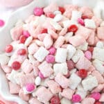 pink and white strawberries and cream puppy chow in white ruffle bowl with m&ms in background