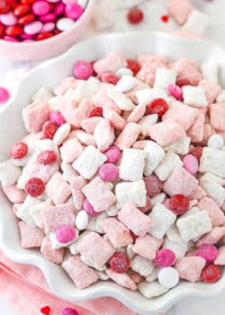 pink and white strawberries and cream puppy chow in white ruffle bowl with m&ms in background
