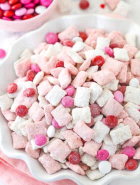 pink and white strawberries and cream puppy chow in white ruffle bowl with m&ms in background