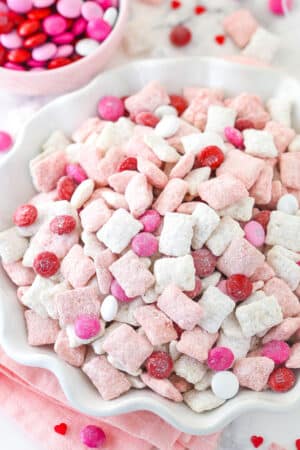 pink and white strawberries and cream puppy chow in white ruffle bowl with m&ms in background