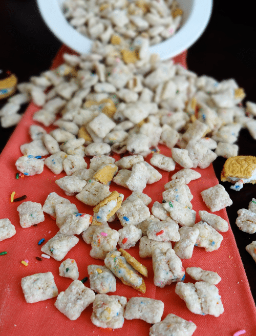 Birthday Cake Puppy Chow spilled out onto a red surface with pieces of Golden Birthday Cake Oreos