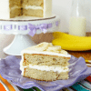 A piece of banana cake on a plate with the remaining cake, a glass of milk and a banana in the background