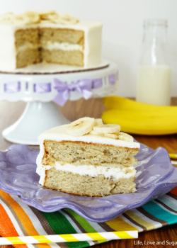 A piece of banana cake on a plate with the remaining cake, a glass of milk and a banana in the background