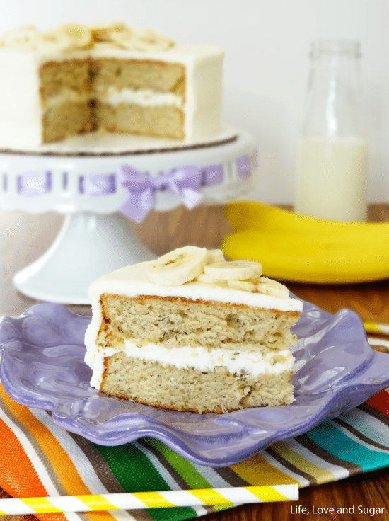 A piece of banana cake on a plate with the remaining cake, a glass of milk and a banana in the background