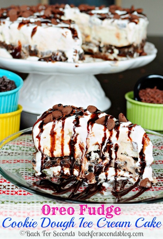 A slice of Oreo Fudge Cookie Dough Ice Cream Cake drizzled with fudge on a small clear plate. 