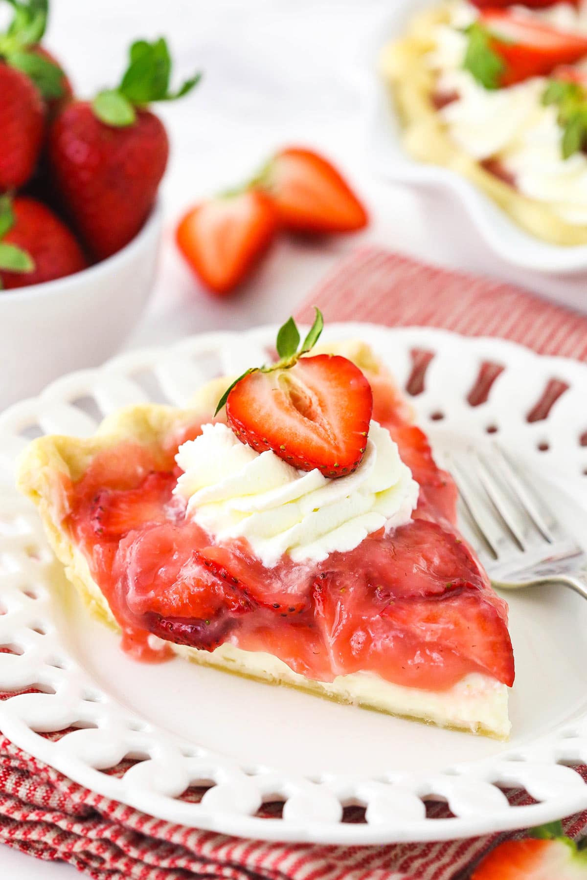 A close-up shot of a slice of strawberry cream pie on a dessert plate with a bowl of fresh berries in the background