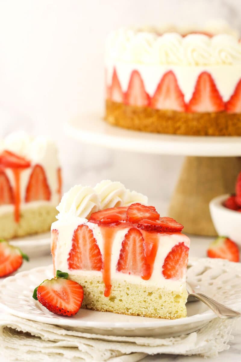 slice of cheesecake on white plate with full cheesecake on cake stand in background