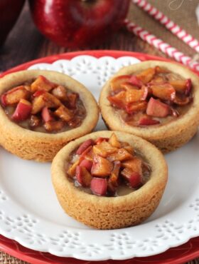 Three apple pie cookies on a white plate with an apple and two straws in the background