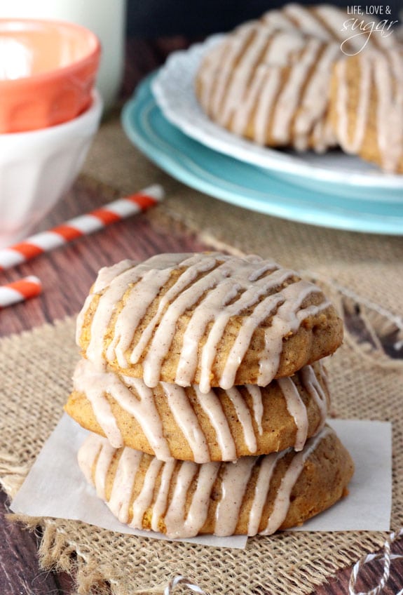 A stack of three glazed pumpkin cookies on a table with a plate holding more cookies behind it.
