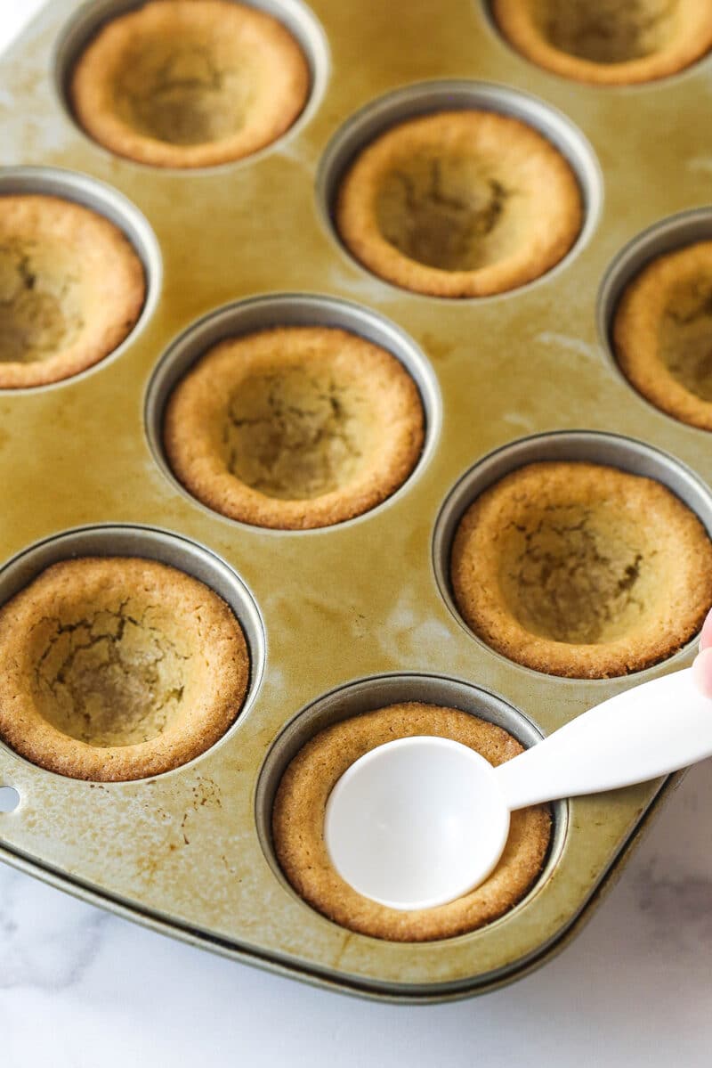 baked cookie cups in cupcake pan with a measuring spoon pushing the center down on one.
