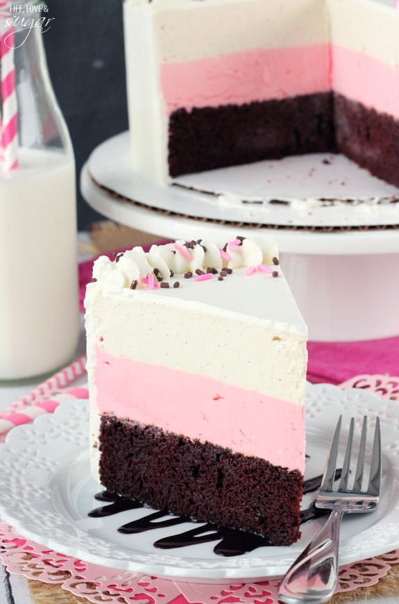 A piece of Neapolitan ice cream cake on a plate with the rest of the cake in the background