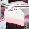 A slice of chocolate, strawberry and vanilla cake on a plate with the remaining cake on a cake stand behind it