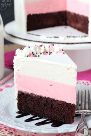 A slice of chocolate, strawberry and vanilla cake on a plate with the remaining cake on a cake stand behind it