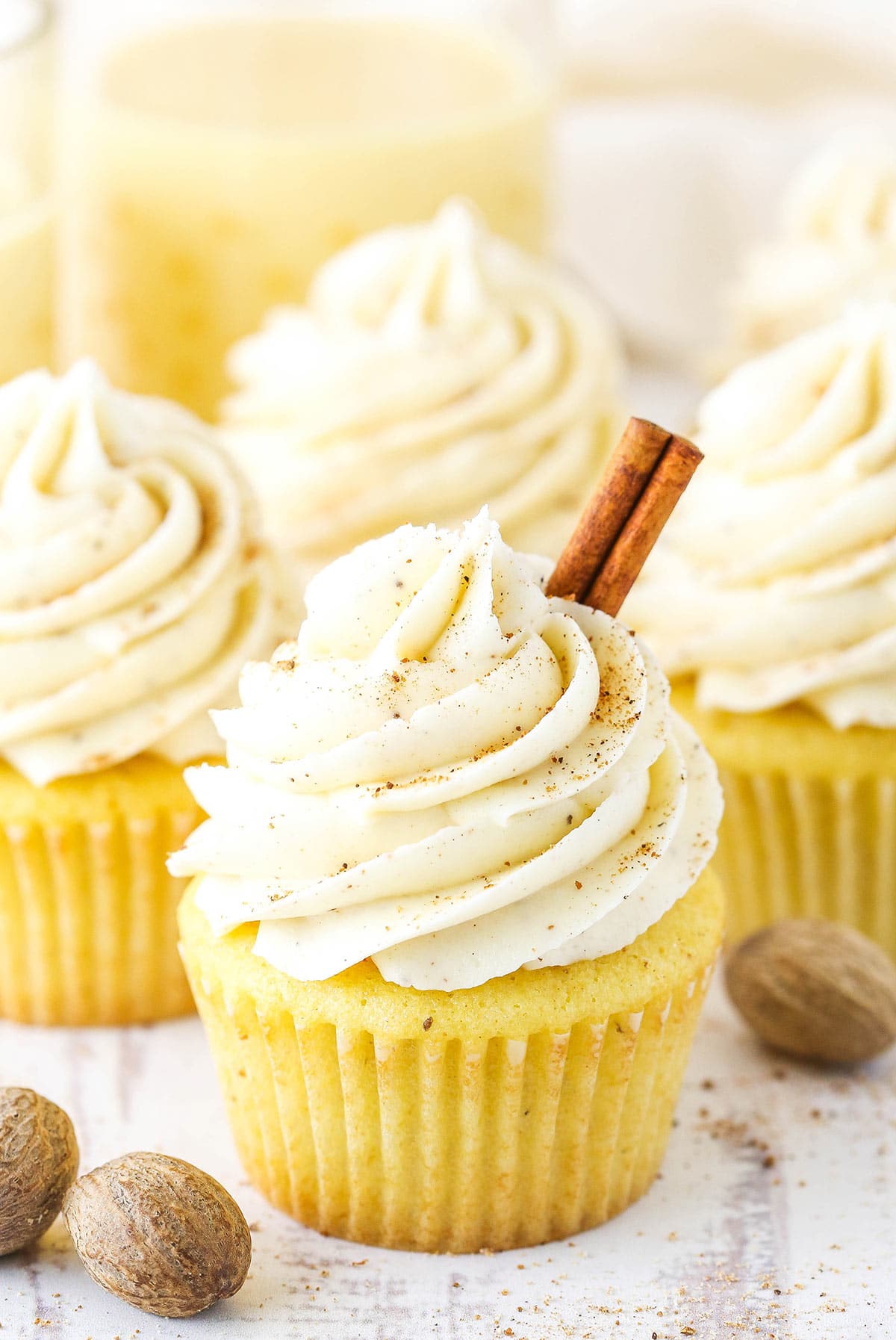 Four eggnog cupcakes on a kitchen countertop with a glass of homemade eggnog behind them