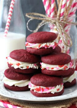 Red Velvet Cookie Sandwiches on white plate stacked with pink, red, white sprinkles