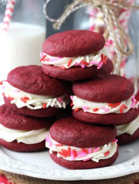 Red Velvet Cookie Sandwiches on white plate stacked with pink, red, white sprinkles