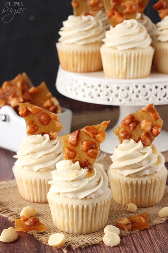 A bunch of cupcakes with icing and macadamia brittle on top surrounded by a cake stand and more nuts