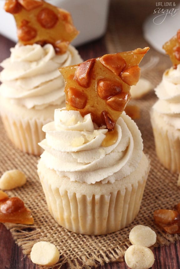 Close up of a Macadamia Brittle Cupcake with icing on top surrounded by more cupcakes and nuts