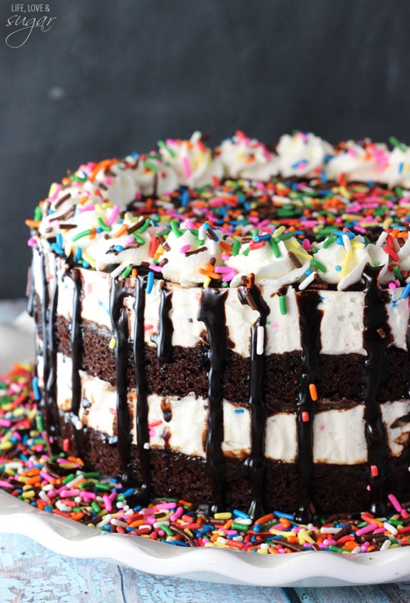 A cake batter fudge brownie Ice cream cake sitting on a dessert stand with rainbow sprinkles on top of the cake