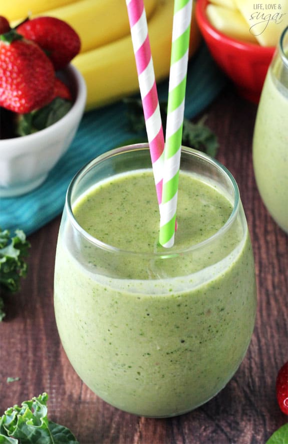 A glass cup on a wood table is filled with a green smoothie and two colorful straws 