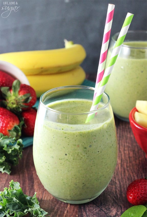 A Green Smoothie in a glass surrounded by spinach, kale, pineapple, strawberry, and bananas