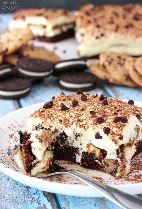A square of layered tiramisu on a small plate, with one bite cut off with a fork on the side of the plate. Oreos and chocolate chip cookies are in the background with the rest of the dessert.