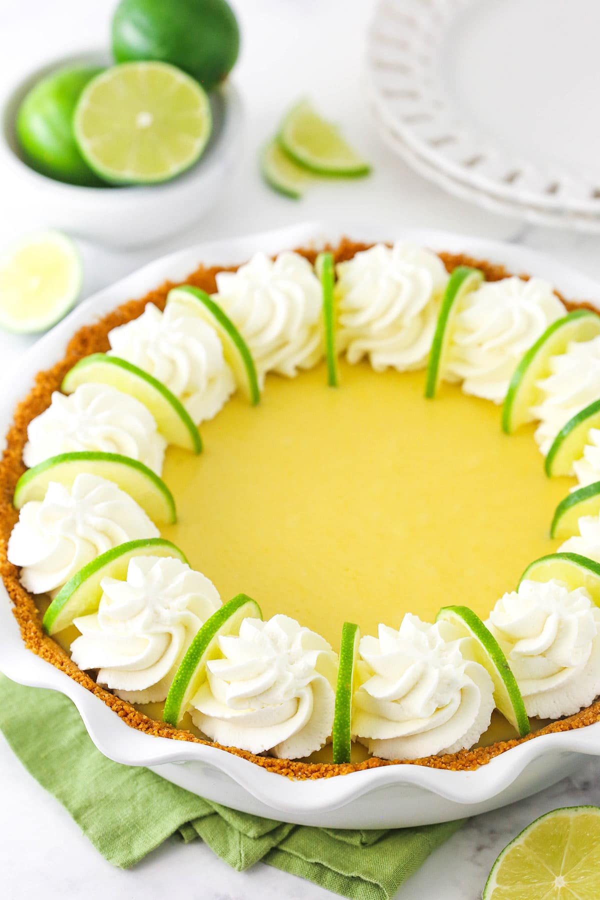 This easy Key Lime Pie recipe is seriously the best! Buttery graham cracker crust with creamy key lime filling and topped with whipped cream. A homemade citrus pie inside of a pie dish with a green kitchen towel underneath the dish and swirls of whipped cream on top of the pie filling
