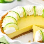 A homemade key lime pie inside of a pie dish with one slice missing and a bowl of fresh limes in the background