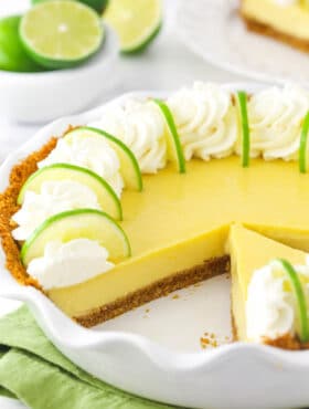 A homemade key lime pie inside of a pie dish with one slice missing and a bowl of fresh limes in the background