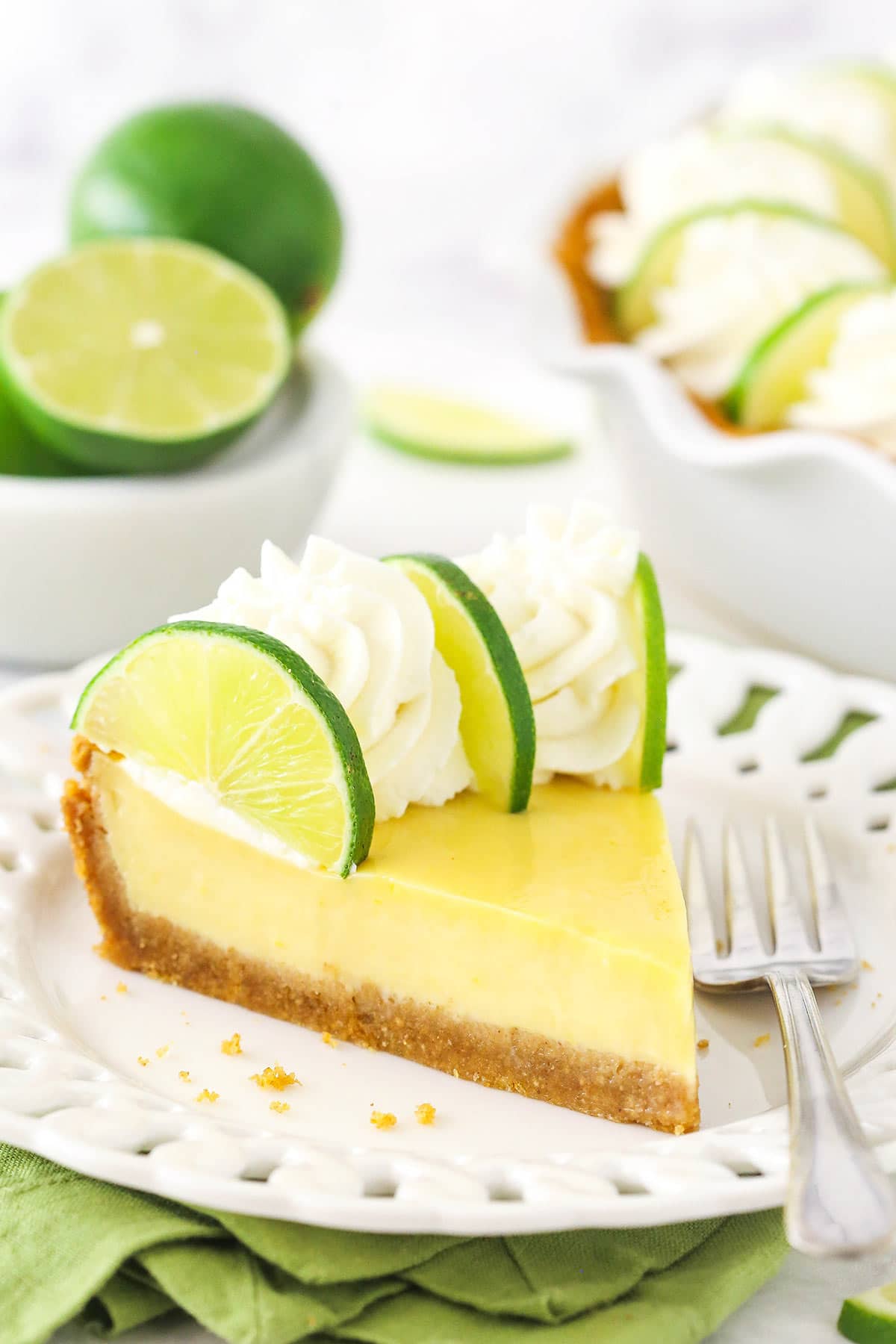 A piece of key lime pie on a plate with a bowl of fresh limes and the rest of the pie in the background