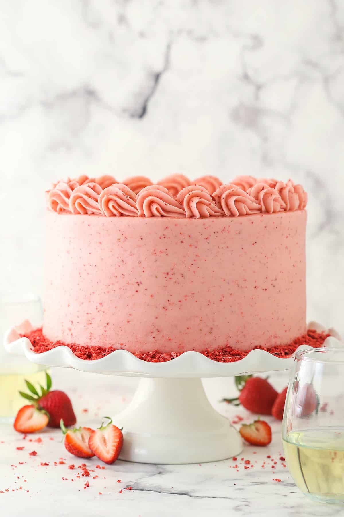 fully frosted strawberry moscato cake on ruffled white cake stand with sliced strawberries around the base