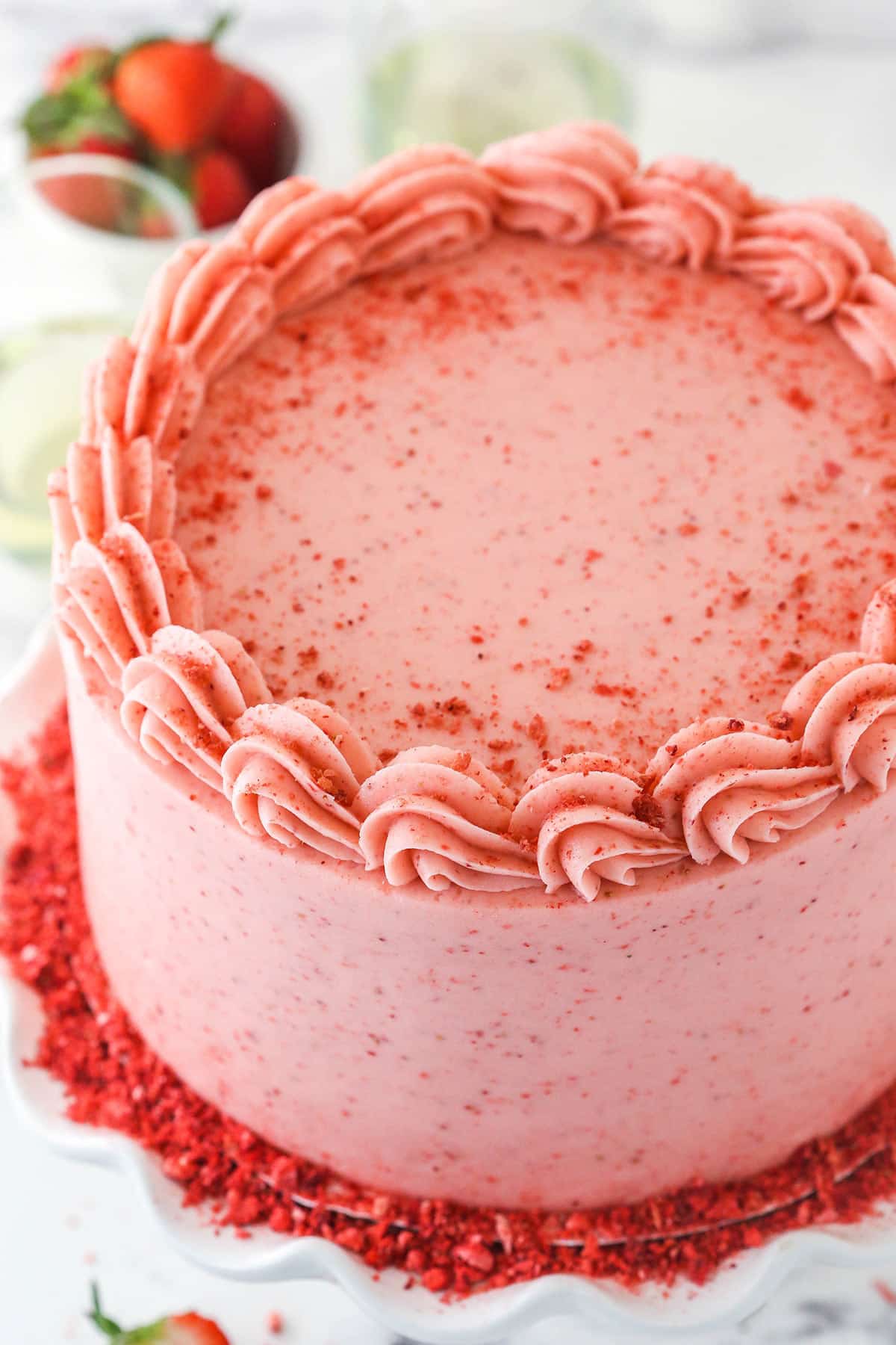 fully frosted strawberry moscato cake on ruffled white cake stand with a bowl of strawberries in the background