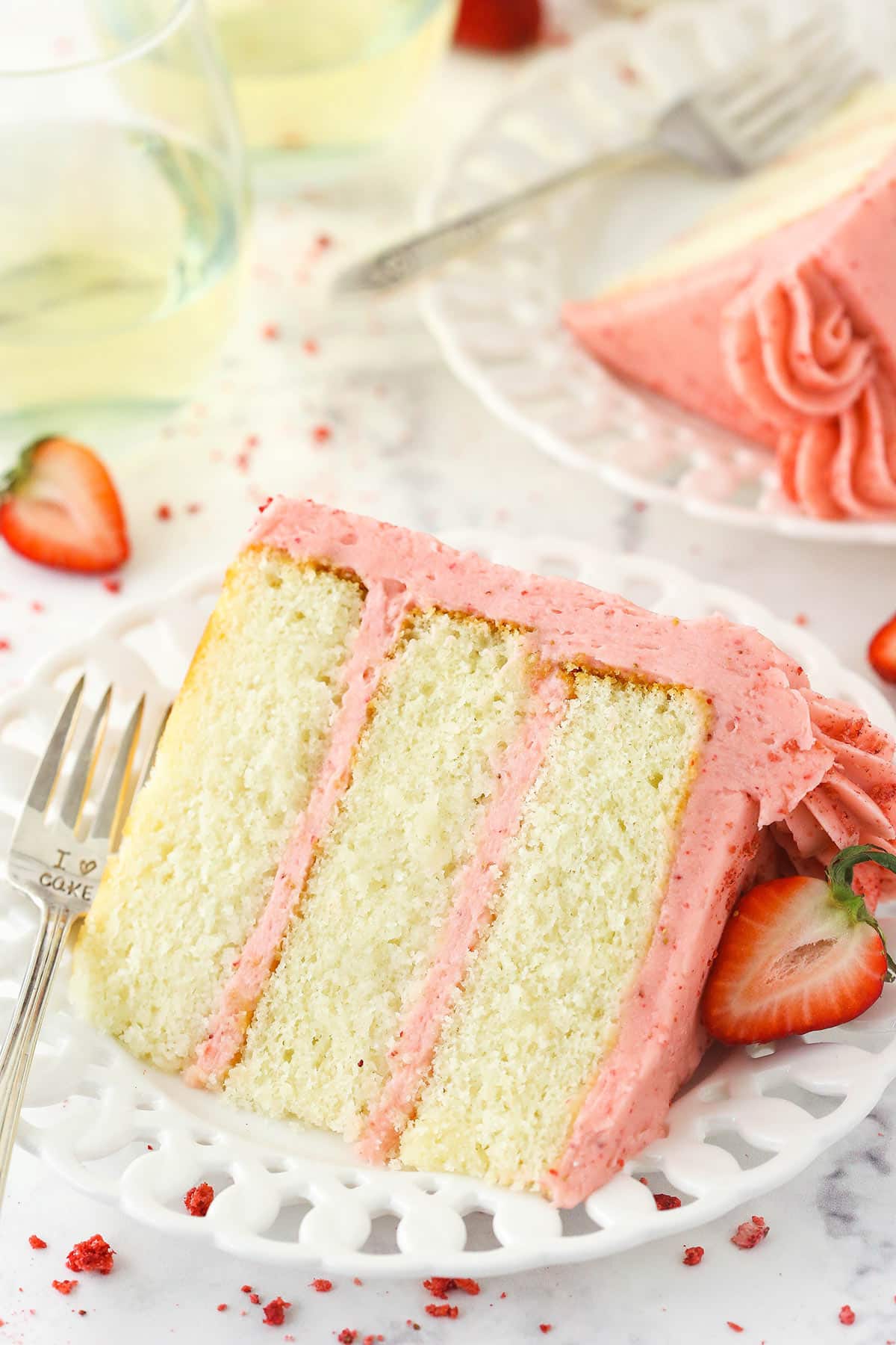 A slice of strawberry moscato cake on a white plate with another slice and some wine glasses in the background.