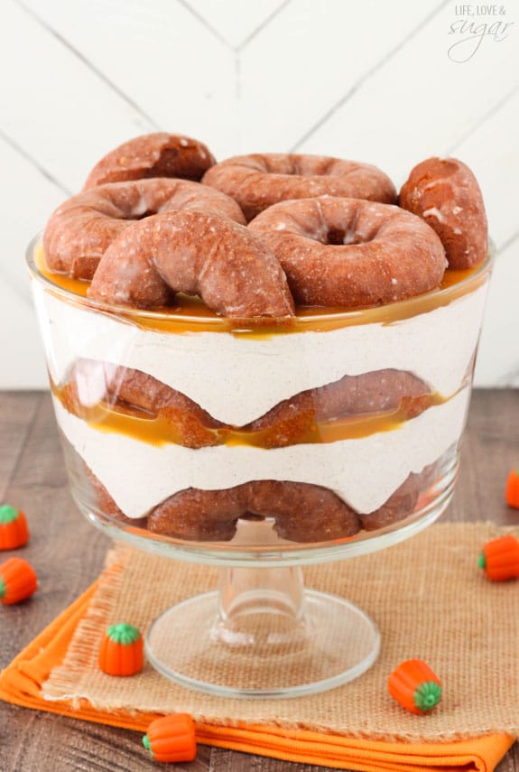 Pumpkin Spice Donut Cheesecake Trifle with layers of pumpkin spice donuts, no bake cheesecake and caramel sauce in a trifle bowl on a burlap and orange cloth