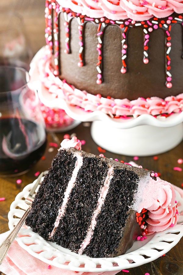 A slice of chocolate cake on a plate in front of the rest of the cake on a stand.