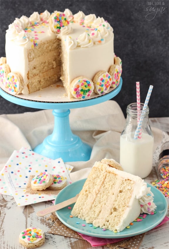 BAILEYS Frosted Vanilla Cookie Layer Cake slice on a blue plate with a glass of milk in front of the whole cake on a blue cake stand
