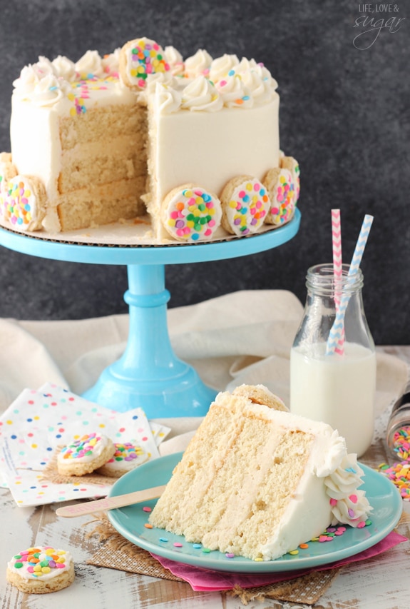 BAILEYS Frosted Vanilla Cookie Layer Cake slice on a blue plate with a glass of milk in front of the whole cake on a blue cake stand