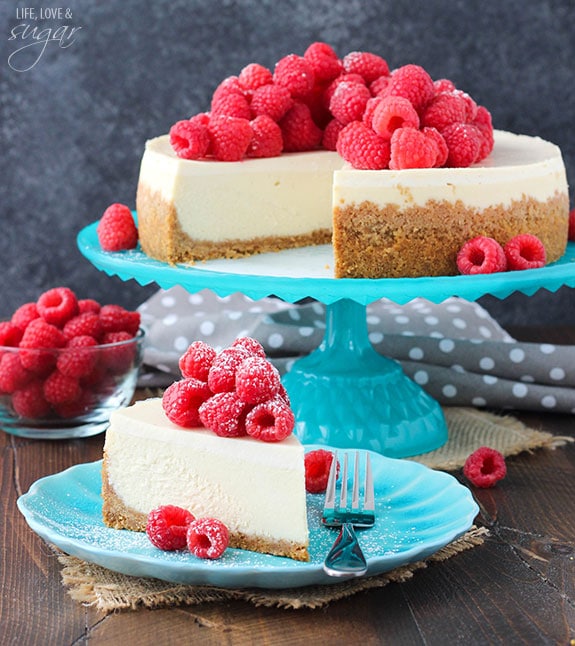 A slice of cheesecake topped with raspberries on a plate in front of the whole cake on a cake stand
