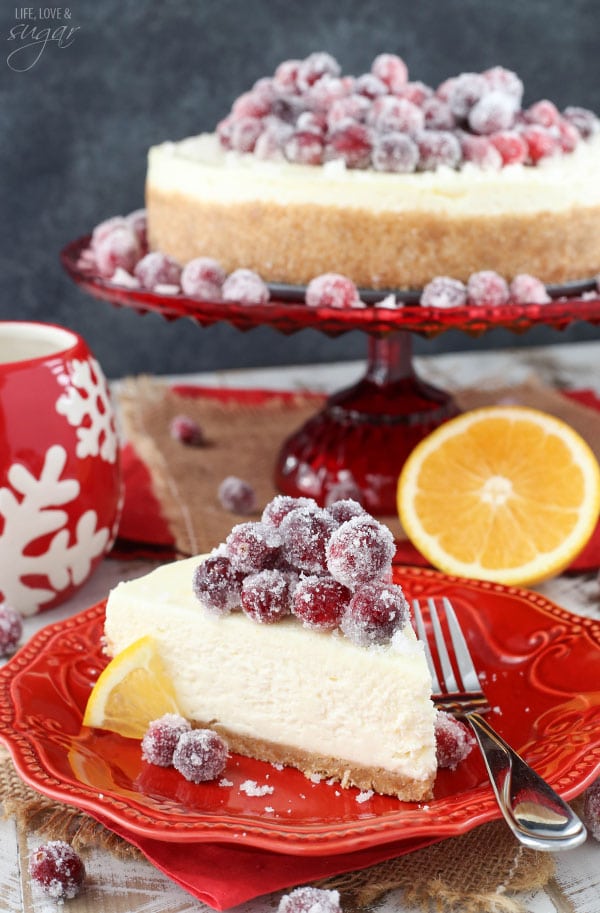 No Bake Sparkling Cranberry Orange Cheesecake slice on a red plate in front of the whole cake on a cake stand