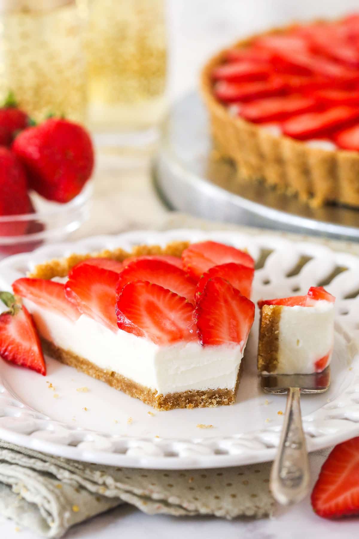 A wafer crust surrounds a creamy champagne mascarpone tart filling, topped with juicy strawberry slices, to create the best strawberry tart! slice of strawberry champagne tart on a white plate with a bite taken