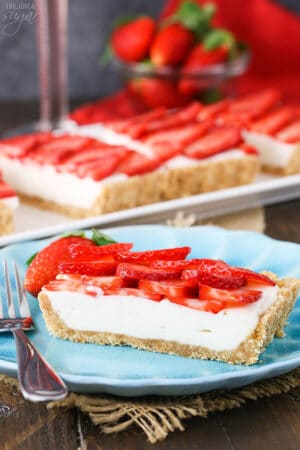 A serving of strawberry tart on a dessert plate with the remaining tart and a bowl of fresh berries in the background