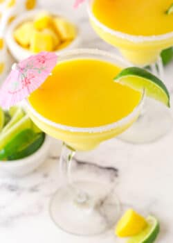mango margarita in a clear glass with lime on rim and pink umbrella on marble counter