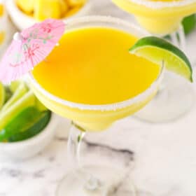 Overhead image of a mango margarita in a clear glass garnished with a lime and a pink umbrella.