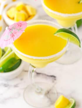 mango margarita in a clear glass with lime on rim and pink umbrella on marble counter