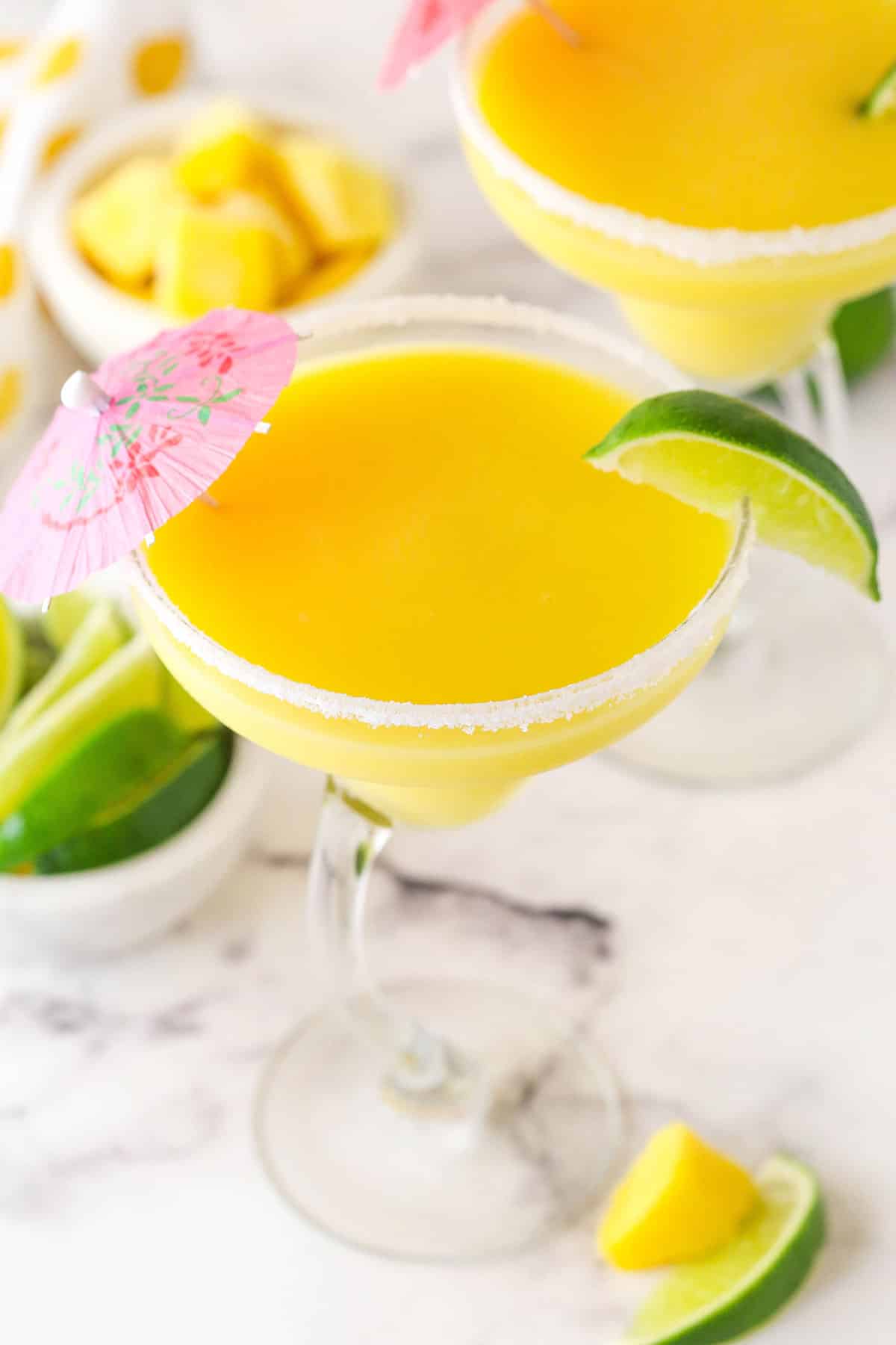 mango margarita in a clear glass with lime on rim and pink umbrella on marble counter