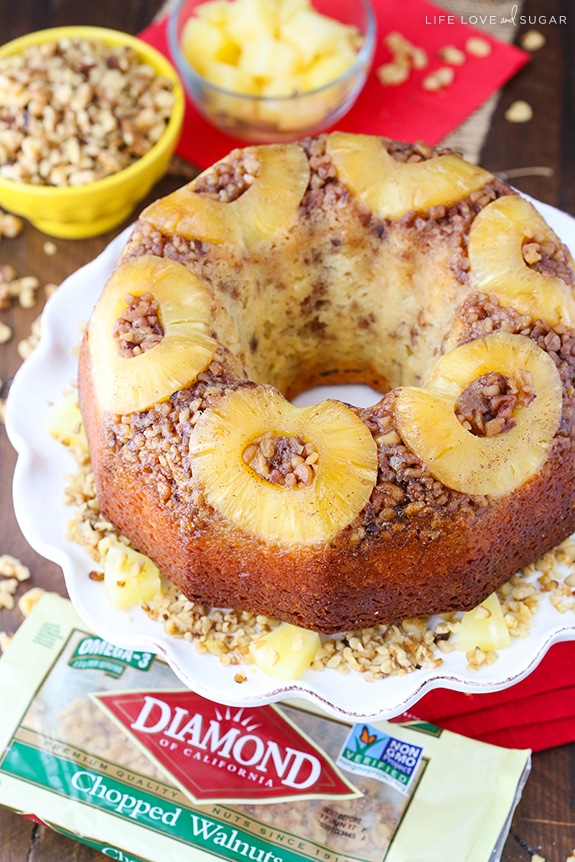 A bundt cake topped with pineapple and nuts sits on a cake stand next to a bag of walnuts