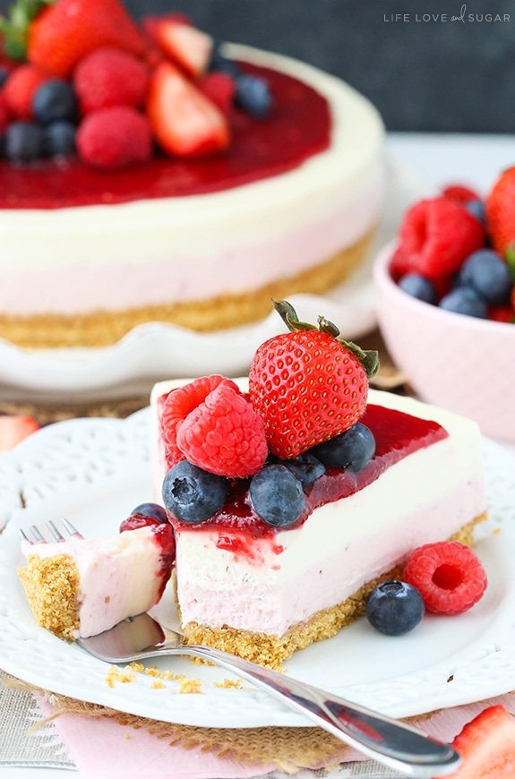 A slice of cheesecake topped with fresh berries is on a plate with a fork removing a piece 